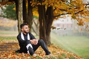 Indian stylish man in black traditional clothes with white scarf posed outdoor against yellow autumn leaves tree. photo