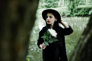 chica sensual todo en negro, labios rojos y sombrero. mujer gótica dramática sostiene una flor de crisantemo blanco a mano contra el lago silencioso. foto