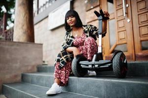 Beautiful african american woman sit near segway or hoverboard. Black girl with dual wheel self balancing electrical scooter. photo