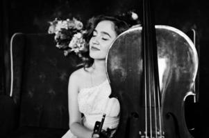 Pretty young gilrl musician in white dress with double bass sitting on brown vintage sofa. photo