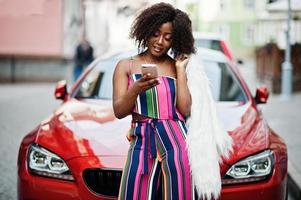 Fashionable african american woman in pink striped jumpsuit with fluffy faux fur coat posed against rich red luxury car with mobile phone at hand. photo