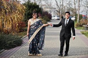 elegante y de moda amigos indios pareja de mujer en sari y hombre en traje caminando al aire libre y tomados de la mano. foto