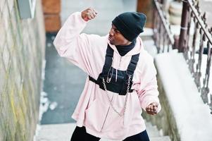 Elegante hombre afroamericano de estilo urbano con capucha rosa posado en el día de invierno muestra los músculos de las manos. foto