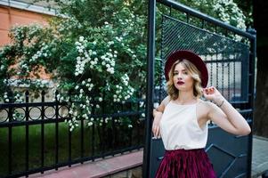 Fashionable and beautiful blonde model girl in stylish red velvet velour skirt, white blouse and  hat, posing outdoor against gates. photo