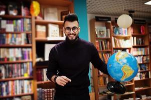 Tall smart arab student man, wear on violet turtleneck and eyeglasses, at library holding earth globe at hands. photo