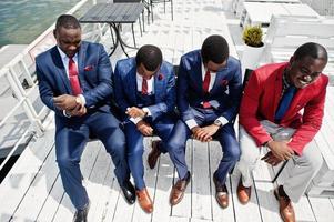 Group of four african american happy succesfull mans at suit. Rich black business mans sitting at chairs and looking at watches. Business time concept. photo