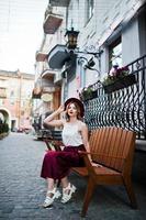 Fashionable and beautiful blonde model girl in stylish red velvet velour skirt, white blouse and hat, posed on bench at city street. photo