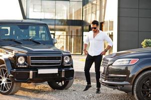 Solid asian man in white shirt and sunglasses posed near black mafia suv cars. photo