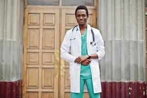 Stylish african american doctor with stethoscope and lab coat posed against door of hospital. photo
