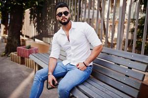 Stylish tall arabian man model in white shirt, jeans and sunglasses posed at street of city. Beard attractive arab guy sitting on bench. photo