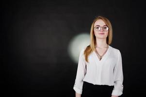 Studio portrait of blonde businesswoman in glasses, white blouse and black skirt against dark background. Successful woman and stylish girl concept. photo