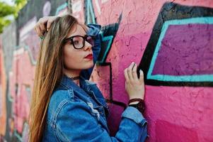 chica hipster casual con estilo en ropa de jeans y gafas contra una gran pared de graffiti. foto