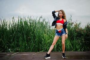 Sexy curly model girl in red top, jeans denim shorts, leather jacket and sneakers posed in the reed. photo