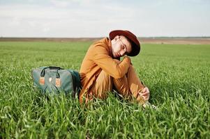 Stylish man in glasses, brown jacket and hat with bag posed on green field. photo