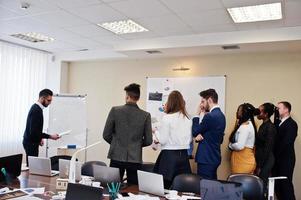 Mixed race business coach presenting report standing near whiteboard pointing on sales statistic shown on diagram and chart teach diverse company members gathered together in conference room. photo