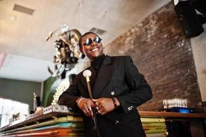 Stylish african american gentleman in elegant black jacket and sunglasses, holding retro walking stick as cane flask or tippling cane with golden diamond ball handle. Rich fashionable afro man. photo