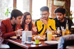 Group of asian friends eating pizza during party at pizzeria. Happy indian people having fun together, eating italian food and sitting on couch. photo