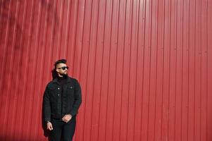 El hombre árabe de moda usa una chaqueta negra de jeans y gafas de sol posadas sobre un fondo de pared de acero rojo. chico modelo árabe elegante y de moda. foto