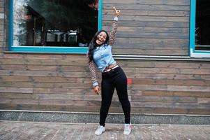 Hipster african american girl wearing jeans shirt with leopard sleeves posing at street against wooden house with windows. photo