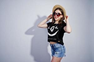 retrato de una chica atractiva en camiseta negra diciendo jajaja, pantalones cortos de mezclilla, sombrero y gafas de sol posando en el estudio. foto