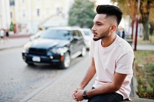 Stylish indian beard man at pink t-shirt sitting against black suv car. India model posed outdoor at streets of city. photo