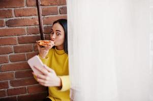 Funny brunette girl in yellow sweater eating pizza at restaurant and making selfie. photo