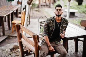 Awesome beautiful tall ararbian beard macho man in glasses and military jacket sitting outdoor wooden table of restaurant. photo