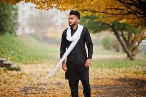 Indian stylish man in black traditional clothes with white scarf posed outdoor against yellow autumn leaves tree. photo