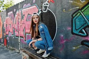 Stylish casual hipster girl in cap and jeans wear listening music from headphones of mobile phone against large graffiti wall with bomb. photo