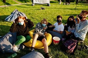 joven grupo multiétnico de personas viendo películas en poof en el cine al aire libre usando una máscara durante la cuarentena del coronavirus covid. foto