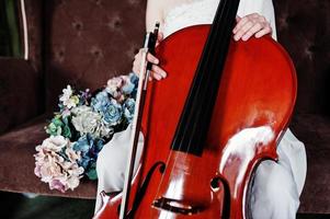 primer plano de un músico gilrl vestido de blanco con contrabajo sentado en un sofá vintage marrón. foto