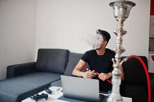 hombre indio guapo y de moda en negro sentado en la habitación, fumando narguile, trabajando en la computadora portátil. foto