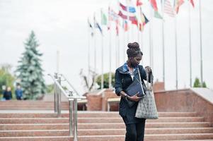 African student female posed with backpack and school items on yard of university, against flags of different countries. photo