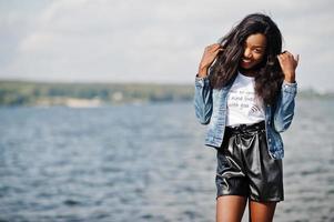 African american dark skinned slim model posed in a black shorts and jeans jacket against sea side. photo