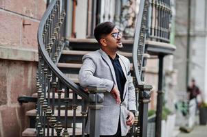 hombre indio elegante con bindi en la frente y anteojos, traje puesto al aire libre contra escaleras de hierro. foto