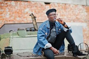 African american man in jeans jacket, beret and eyeglasses, smoking cigar and posed against btr military armored vehicle. photo