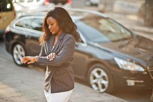 éxito elegante mujer afroamericana en chaqueta contra coche negro mirando sus relojes.