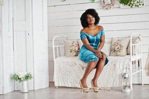 Afican american women in brilliant glitter sequins turqoise dress sitting on vintage couch at white room. photo