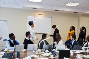 Mixed race business coach presenting report standing near whiteboard pointing on sales statistic shown on diagram and chart teach diverse company members gathered together in conference room. photo