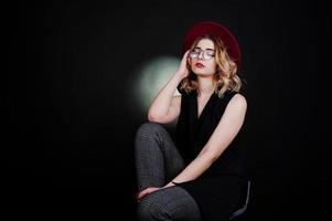 retrato de estudio de una chica rubia con ropa negra, sombrero rojo y anteojos contra un fondo oscuro. foto