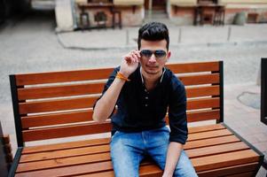 Stylish indian model man in casual clothes, black shirt and sunglasses posed outdoor at street of India and sitting on bench. photo