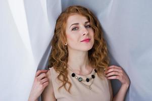 Curly hair girl on beige dress posed at white curtains. photo