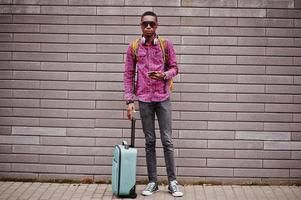 African american man in checkered shirt, sunglasses and earphones with suitcase and backpack. Black man traveler against wall holding mobile phone. photo