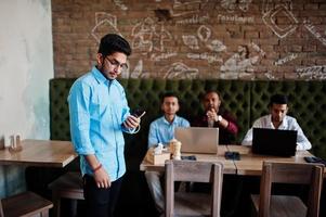 Group of four south asian men's posed at business meeting in cafe. Indians work with laptops together using various gadgets, having conversation. Indian man with mobile phone. photo