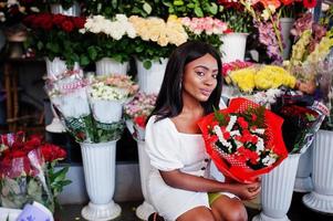 Beautiful african american girl in tender white dress with bouquet flowers in hands standing against floral background in flower shop.Black female florist. photo