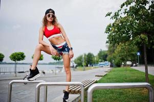 Sexy curly model girl in red top, jeans denim shorts, cup sunglasses and sneakers posed against steel rails. photo