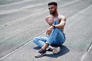 Handsome sexy african american bare torso man at jeans overalls sitting at stadium racecourse. Fashionable black man portrait. photo