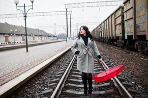 Brunette girl in gray coat with red umbrella in railway station. photo