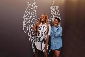 Two african womans in stylish casual clothes posing against angel wings wall. photo