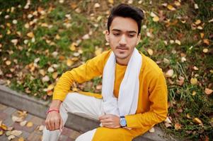Indian stylish man in yellow traditional clothes with white scarf posed outdoor against autumn leaves. photo